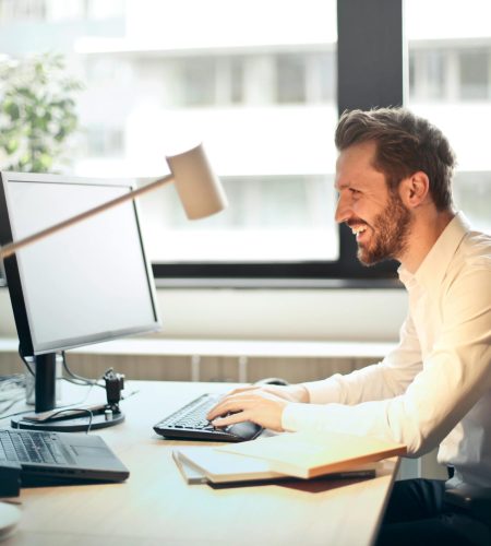 A man smiling while working at an office desk with a computer and natural daylight streaming in through large windows.